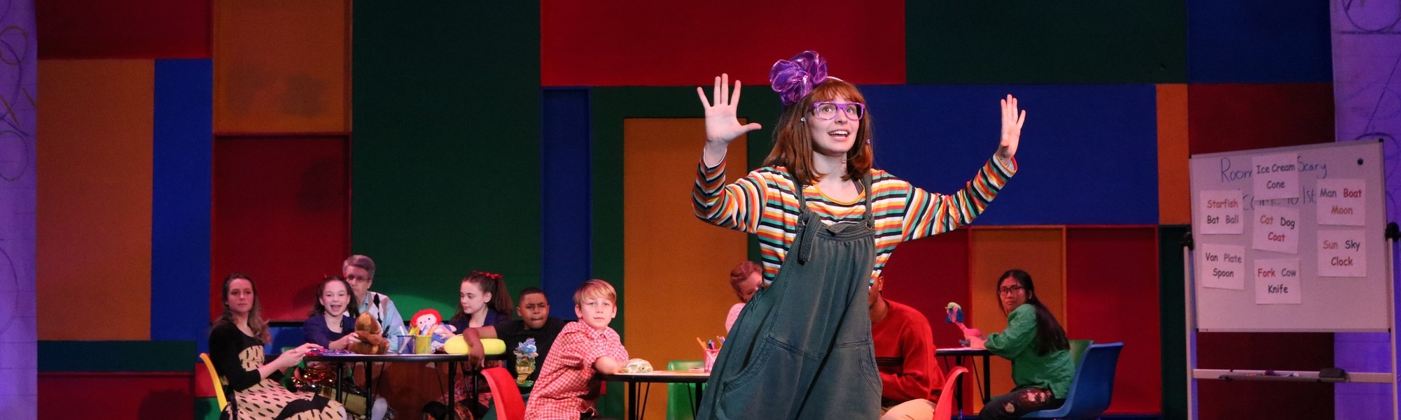 Young female actress with purple glasses and red hair looking excited with her hands up in front of a colorful set and other children sitting at tables.