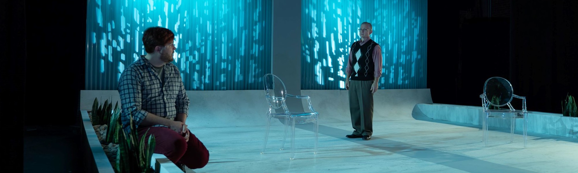Two actors on a barren stage with blue lights in the background staring tensely at each other.