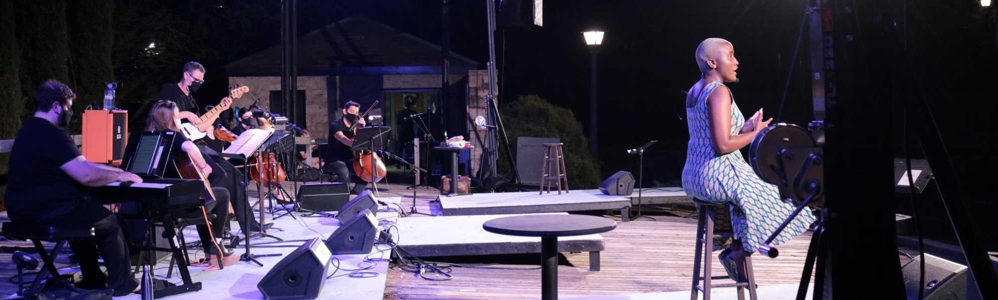 Side view of an outdoor stage with several musicians in black playing their instruments and a dark skinned female actress sitting on a stool and singing.
