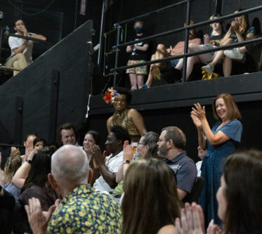 An African-American person with bright flowers in her hair stands up smiling as the crowd seated in the theater applauds.